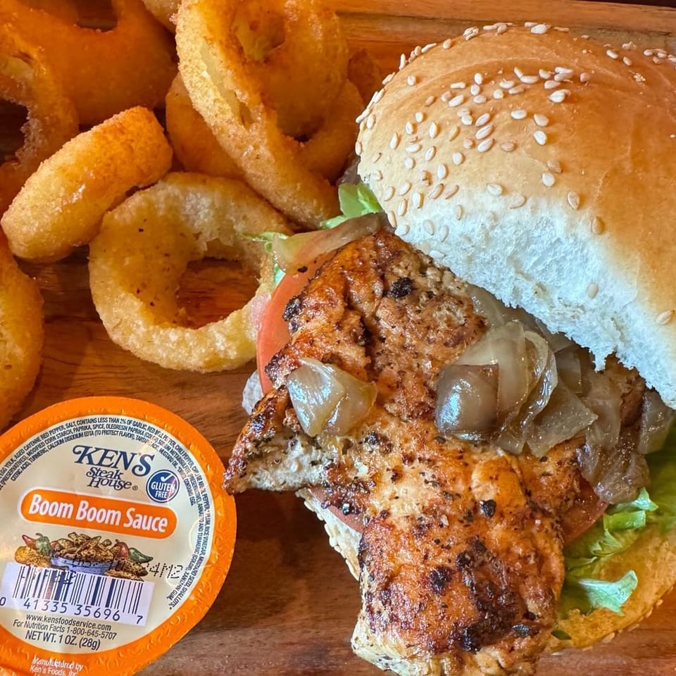 A grilled chicken sandwich with caramelized onions, lettuce, and tomato, served with onion rings and Ken's Boom Boom Sauce.