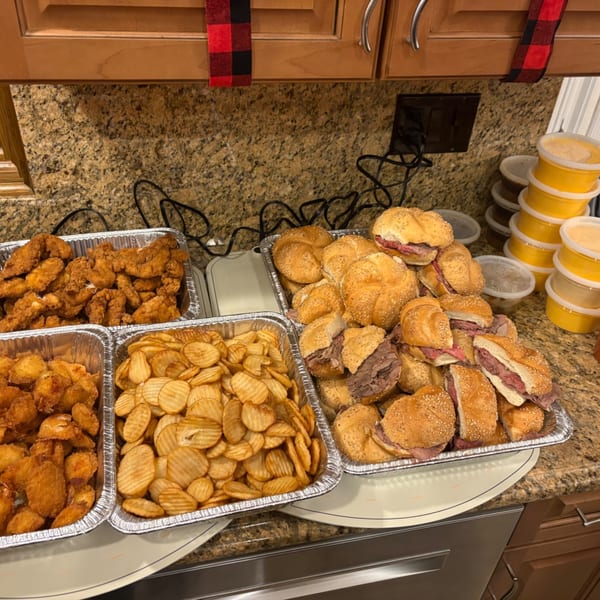 Catering trays of fried chicken, waffle chips, and roast beef sandwiches on a granite countertop.