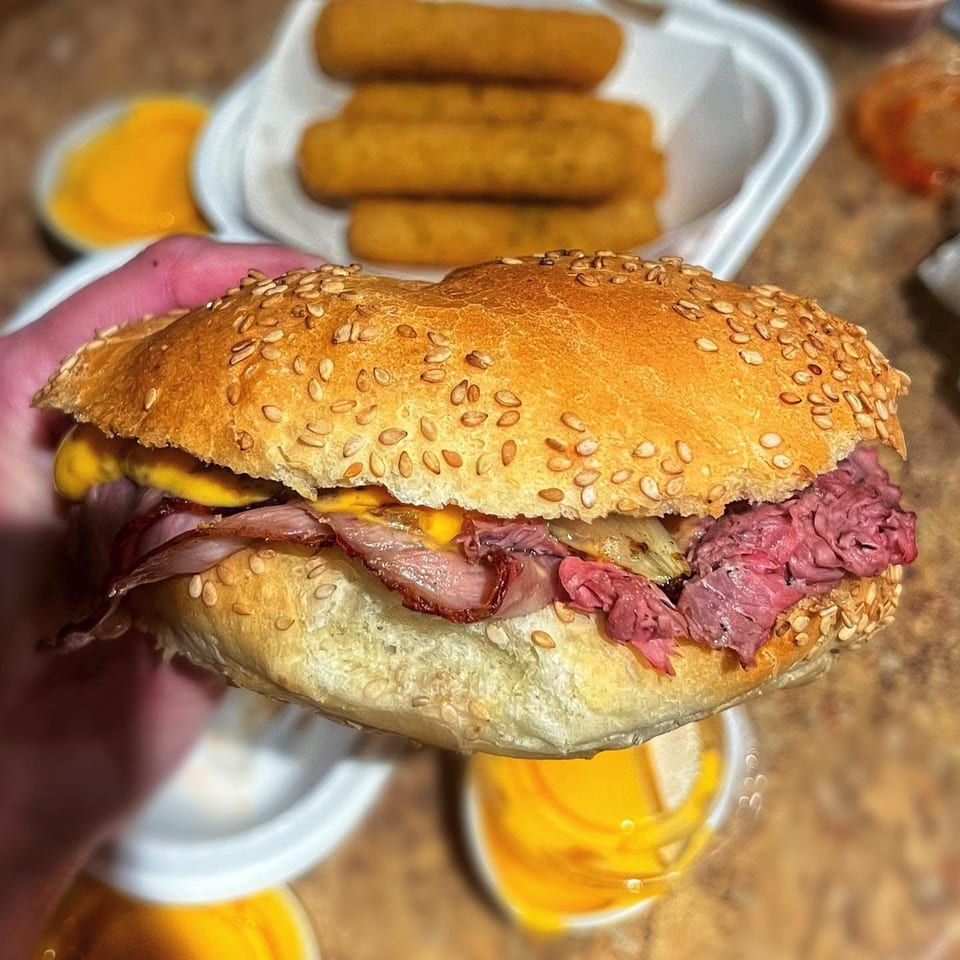 Hand holding a pastrami and cheese sandwich on a sesame seed bun, with mozzarella sticks in the background.